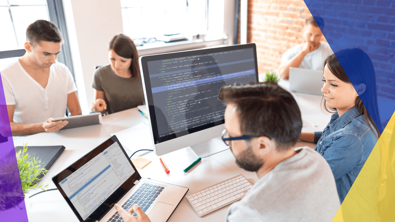 Five developers at a shared white desk with laptops and a monitor of code, a bearded man in glasses typing up front.