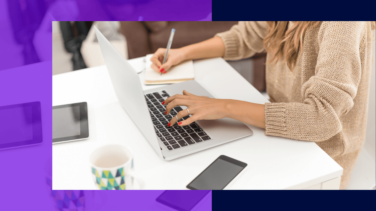 Woman in a beige knit sweater typing on a laptop and writing in a notebook at a white desk beside a tablet and mug.