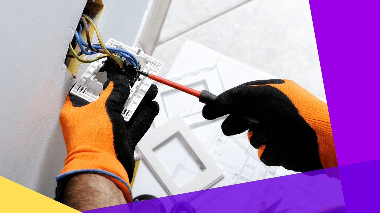 Electrician in orange work gloves using a red screwdriver on wires in an open wall socket, floor plans in the background.