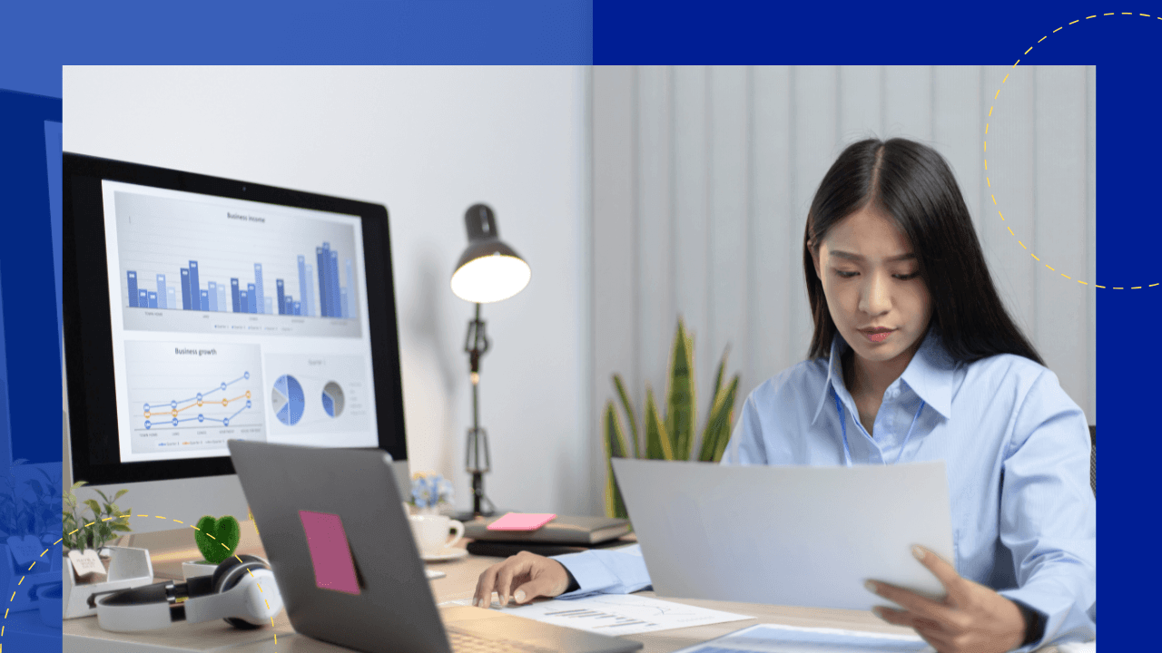 Woman in a light blue shirt reviewing a printed document at a desk beside a laptop and a monitor showing business charts.
