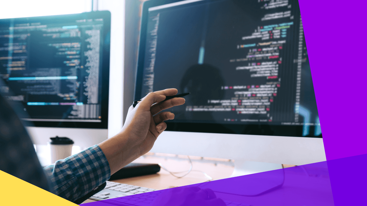 Hand holding a pen and pointing at lines of code on a desktop monitor, a second screen of code and a coffee cup beside it.