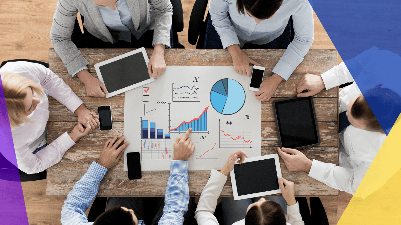 Overhead view of a team around a wooden table with tablets and phones, pointing at a printed sheet of charts and graphs.